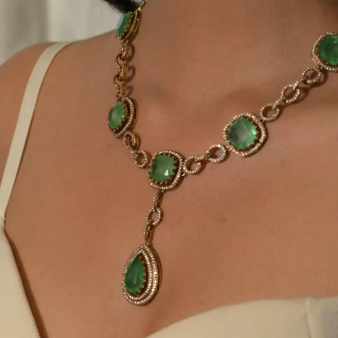 Elegant emerald and diamond necklace with gold chain, featuring teardrop pendant and round gemstones.