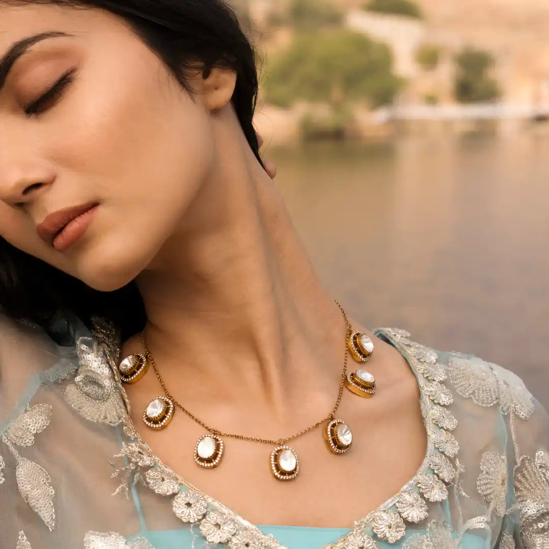 Elegant gold necklace with white gemstones, worn by woman in embroidered blue attire near water