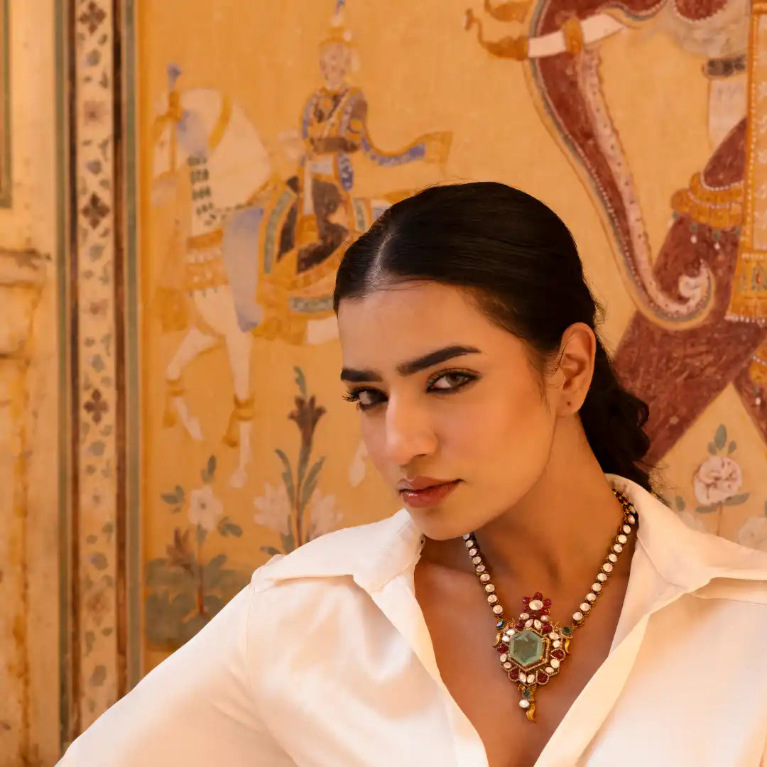 Woman wearing ornate gemstone necklace with green center stone, styled against traditional mural backdrop