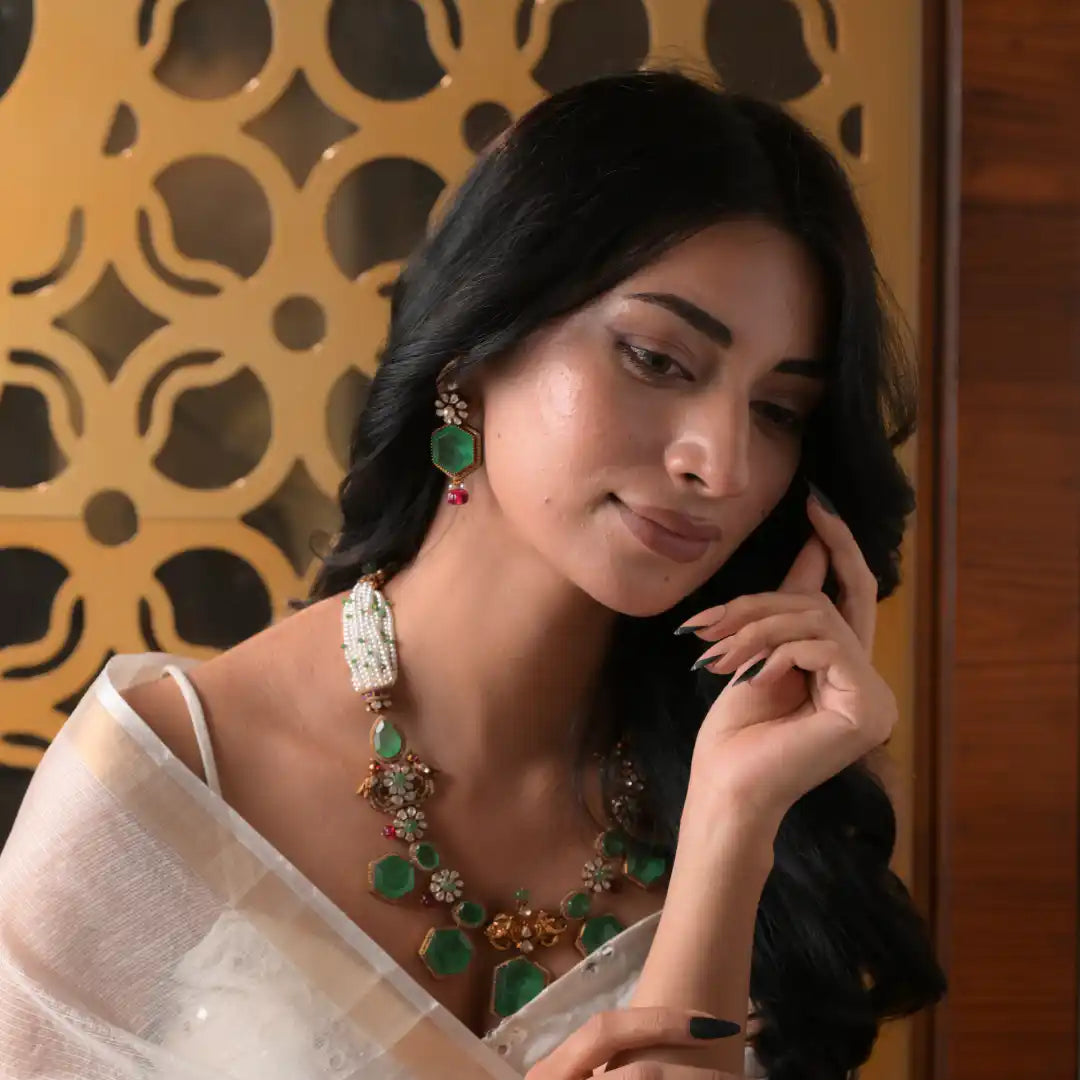 Woman wearing elegant green gemstone necklace and matching earrings, styled with white saree against ornate gold backdrop