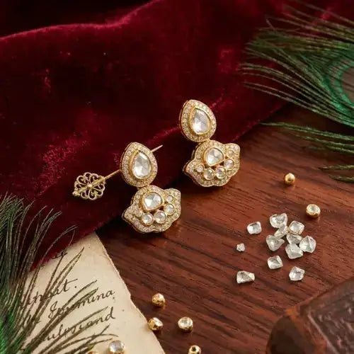 Elegant gold earrings with white stones, displayed on velvet with peacock feathers and loose gemstones